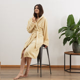 Frau trägt LUMALAND Luxury Mikrofaser Bademantel mit Kapuze für Damen und Herren - Creme - Größe S, sitzend auf Hocker, gemütlich