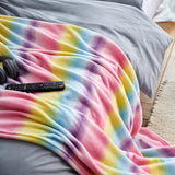LUMALAND Kuscheldecke für Kinder 100x150 cm in buntem Regenbogen-Multicolour-Design aus weichem Coral-Fleece auf grauem Bett.