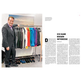 Ein Mann im Anzug steht neben einem Kleiderständer, unter dem bunte Jacken und Schuhe stehen.