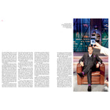 Ein Mann im Anzug sitzt auf einem braunen Stuhl, im Vordergrund hält jemand ein Telefon hoch und macht ein Foto.