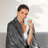 Frau entspannt auf Sofa mit MAXXMEE Heizdecke 180 x 130cm, grau, wärmt sich mit einer Tasse Tee und Fernbedienung.