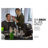 Eine Person wird in einem Studio von einem Künstler geschminkt. Auf dem Tisch stehen Make-up-Utensilien und grüne Becher.