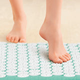 Nackte Füße treten auf eine weiße, mit Stacheln versehene Akupressurmatte auf einem hellen Boden.