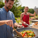 Mann nutzt LANDMANN Grillwok aus emailliertem Gusseisen für vielseitiges Grillen auf Gas- oder Holzkohlegrill im Garten.