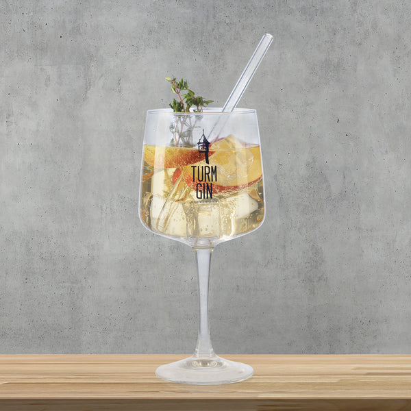 TURM GIN Copa Glas mit Logo und Schriftzug - 720 ml - stilvolles Gin-Glas mit Eis und Kräutern auf Holztisch vor grauem Hintergrund.